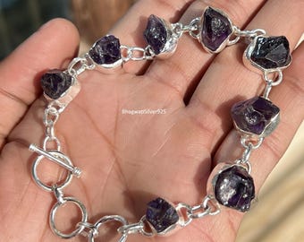 May include: A silver bracelet featuring raw, dark purple amethyst stones. The bracelet is composed of individual stones connected by silver links and a toggle clasp. The text "BhagwatiSilver925" is visible.