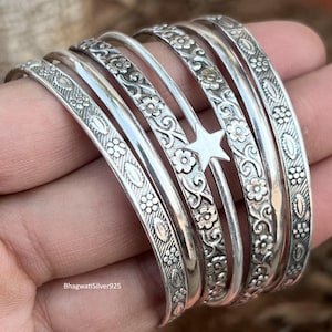 May include: A set of silver bangles featuring floral and star motifs. The bangles display detailed patterns and textures, with a combination of smooth and ornate surfaces. The jewellery is shown in a hand.