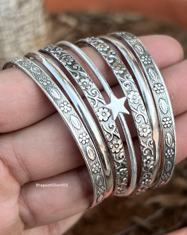 May include: A set of silver bangles featuring floral and star motifs. The bangles display detailed patterns and textures, with a combination of smooth and ornate surfaces. The jewellery is shown in a hand.