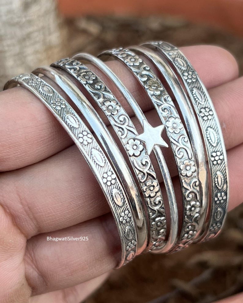 May include: A set of silver bangles featuring floral and star motifs. The bangles are displayed together, highlighting the detailed patterns and textures. The designs include flowers, leaves, and a star, creating a refined and elegant appearance.