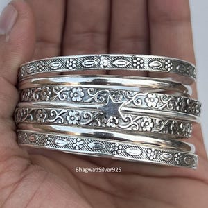 May include: Several silver bangles with detailed floral and leaf designs. The bangles are held in a hand, highlighting the engraved patterns and the polished, reflective surface. The text "BhagwatiSilver925" is visible.