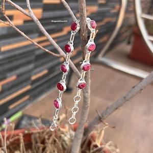 May include: A silver chain bracelet featuring multiple round, deep red ruby gemstones set in individual metal bezels. The bracelet includes a lobster claw clasp and a series of circular links for adjustment. The text BhagwatiSilver925 is visible.