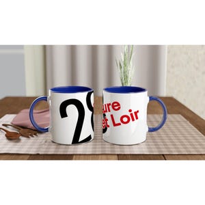 May include: Two white ceramic mugs with blue handles and interiors. One mug displays the number "28" in black, while the other reads "ture et Loir" in red. The mugs are on a checkered tablecloth.