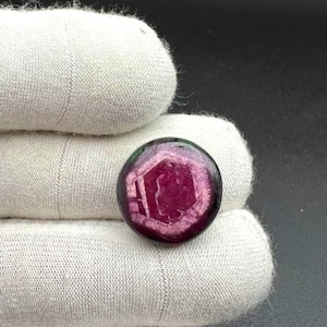 Könnte beinhalten: Ein runder, polierter Edelstein mit einer Farbpalette aus tiefem Magenta und Dunkelviolett. Der Stein zeigt ein ausgeprägtes, helleres sechseckiges Muster in seiner Mitte, das natürliche Variationen in der Oberflächenstruktur aufweist.