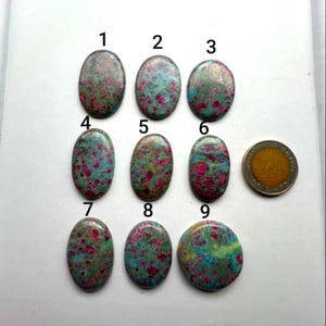 Op de afbeelding: Negen ovaalvormige robijn-zoisiet cabochons gerangschikt in drie rijen, elk gelabeld met een zwart nummer van één tot negen. De stenen hebben een gevlekt patroon van levendig magenta, blauwgroen en groen. Een goudkleurige munt ligt naast steen nummer zes.