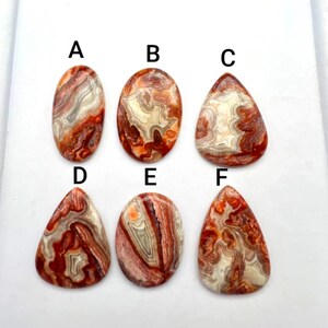 以下が含まれることがあります： AからFのラベルが付いた6つの研磨されたカボション。赤、オレンジ、白、クリーム色の渦巻き模様が特徴です。石は2列に配置されており、楕円形と涙滴形の形状で、複雑な縞模様と層状の鉱物テクスチャを示しています。