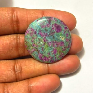 May include: A circular gemstone featuring a mottled pattern of magenta, teal, and green. The polished stone has a smooth surface and a mix of colours, with magenta forming irregular shapes throughout the teal and green base.