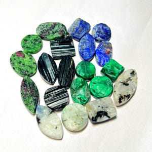 May include: A collection of assorted polished gemstones in various shapes and sizes. The stones include green and red speckled pieces, deep blue lapis lazuli, black striated tourmaline, vibrant green malachite, and off-white stones with black inclusions.