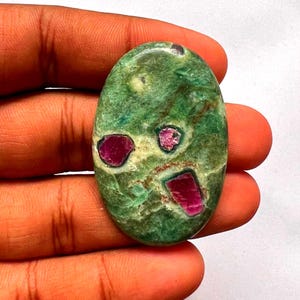 Puede incluir: Una piedra preciosa en forma de cabujón ovalado con una base moteada en verde y blanco, que presenta tres inclusiones incrustadas de color rojo rosado intenso. La superficie de la piedra es pulida y lisa, mostrando texturas naturales.
