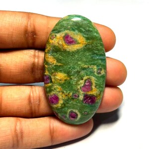 Puede incluir: Una piedra preciosa en forma de cabujón ovalado con una superficie pulida, una base verde moteada, intercalada con distintas inclusiones de color rosa magenta y parches de depósitos minerales amarillo dorado en su cara lisa.