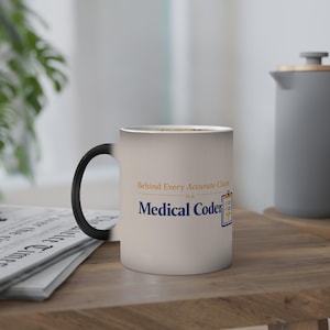Könnte beinhalten: Ein weißer Keramikbecher mit schwarzem Henkel, der den Text "Behind Every Accurate Claim is a Medical Coder" in blauer und goldener Schrift trägt, ergänzt durch eine kleine Grafik eines Klemmbretts mit einem medizinischen Symbol.