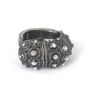 May include: A dark silver-toned ring featuring a textured, rounded surface with a pattern of small, raised bumps and embedded clear, sparkling stones. The metal has an oxidized finish that highlights the intricate, organic-inspired detailing.