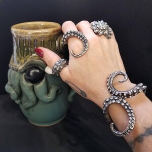 May include: A silver-toned octopus tentacle bracelet, a tentacle ring, a textured band ring, and a cluster ring. Beside these accessories is a green ceramic mug featuring a sculpted octopus face with a prominent black eye and tentacles.