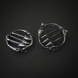 Puede incluir: Un par de pendientes de plata con un diseño circular que presenta tres flechas apuntando hacia afuera. Los pendientes tienen un poste y un cierre de mariposa.