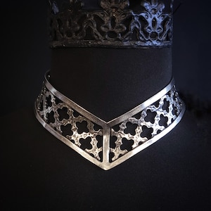 Puede incluir: Collar de metal plateado con un diseño calado de formas de trébol de cuatro hojas repetidas. El collar tiene un frente en forma de V y se exhibe en un maniquí negro. Una corona negra es visible en el fondo.