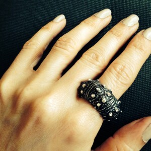May include: A wide, dark-toned metal ring featuring a textured, bumpy surface adorned with numerous small, clear, sparkling stones embedded across the exterior. The band has a substantial, rounded profile that covers a significant portion of the finger.