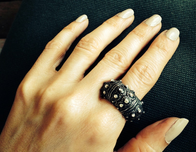May include: A wide, dark-toned metal ring featuring a textured, bumpy surface adorned with numerous small, clear, sparkling stones embedded across the exterior. The band has a substantial, rounded profile that covers a significant portion of the finger.