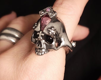 Puede incluir: Un anillo de calavera de plata con una gema rosa en la parte superior. La calavera tiene detalles intrincados y un diseño de corona.