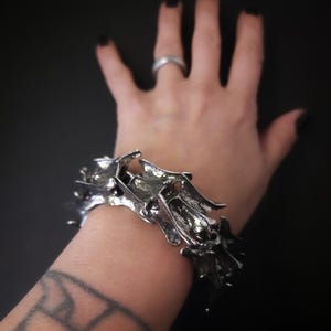 May include: A silver bracelet with a unique, organic, and textured design. The bracelet is made of multiple interconnected pieces that resemble branches or thorns.