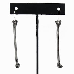 May include: A pair of silver bone-shaped earrings. The earrings are long and slender, with a rounded end. They are hanging from a black earring display stand.
