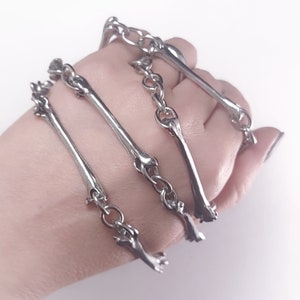 Könnte beinhalten: Silberfarbene Knochen-Anhänger-Armbänder mit Kettengliedern. Die Armbänder sind übereinander am Handgelenk einer Person gelegt.