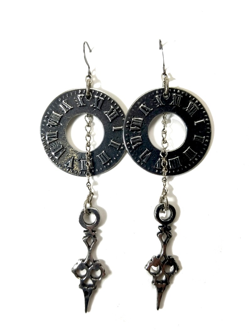May include: Silver clock face earrings with a dangling clock hand design. The clock face has Roman numerals.
