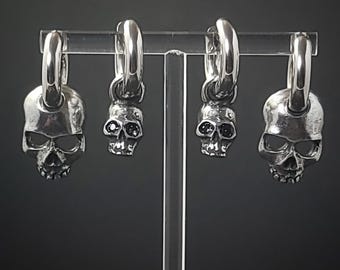 May include: Four silver skull earrings with hoop closures. Two pairs of earrings have black gem eyes. The skulls are detailed with open eye sockets, noses, and teeth. The earrings are displayed on a clear stand against a dark background.