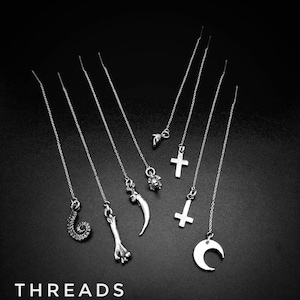 Peut inclure: Boucles d'oreilles pendantes en argent avec différents breloques, dont une croix, un croissant de lune et un os. Elles sont présentées sur fond noir. Le mot "THREADS" est en bas.