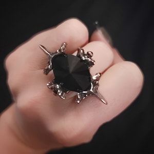 Op de afbeelding: Een zilverkleurige ring met een grote, facetgeslepen zwarte edelsteen. De ring heeft een uniek, getextureerd ontwerp met klauwachtige tanden die de steen vasthouden. De donkere edelsteen contrasteert met het zilvermetaal.