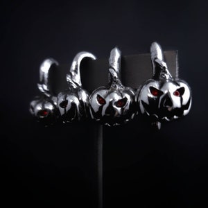 Puede incluir: Cuatro pendientes de calabaza plateados con ojos de piedras preciosas rojas. Los pendientes se muestran en un soporte negro.