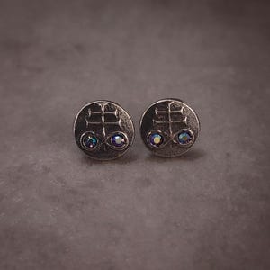 May include: A pair of silver-toned stud earrings. Each earring features a circular design with a symbol and two iridescent, round gemstones. The earrings are displayed on a gray surface.