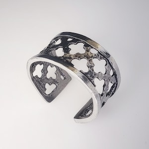 May include: A silver-coloured cuff bracelet featuring an openwork clover design. The bracelet has a wide band with a curved opening. The metal has a slightly oxidised finish, giving it a vintage appearance. The clover cutouts create a decorative pattern.