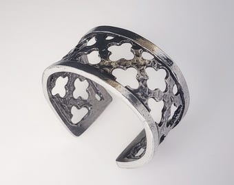 May include: A silver-toned cuff bracelet with an openwork clover design. The bracelet has a wide band with a curved opening. The metal has a slightly oxidized finish, giving it a vintage appearance. The clover cutouts create a decorative pattern.