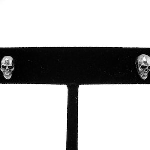 May include: A pair of silver skull stud earrings displayed on a black velvet jewelry stand.