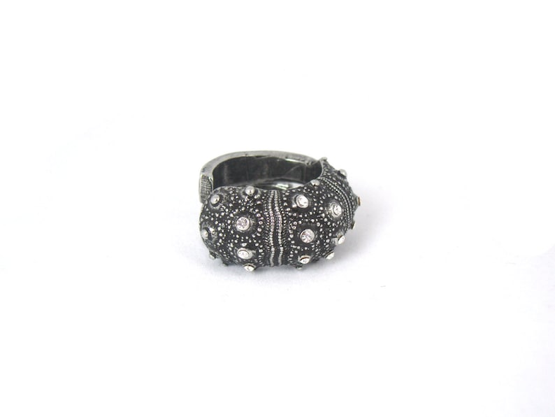 May include: A dark silver-toned ring featuring a textured, rounded surface with a pattern of small, raised bumps and embedded clear, sparkling stones. The metal has an oxidized finish that highlights the intricate, organic-inspired detailing.