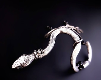 May include: A silver-toned snake bracelet featuring a textured, scale-like surface. The snake head is adorned with a small, intricate collar detail, and the body curves to incorporate smooth, round black beads integrated into the metal structure.