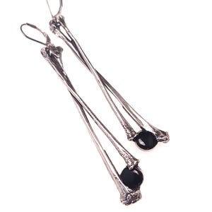 May include: A pair of silver-toned, long dangle earrings. Each earring features a bone-like design with a black, round bead at the bottom. The earrings have a hook closure and are set against a white background.