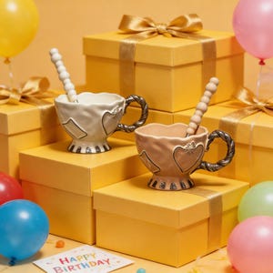 May include: Two decorative teacups with spoons, featuring heart designs and ornate handles, sit atop yellow gift boxes. The teacups are in light beige and peach colours. Balloons and a "Happy Birthday" card are in the foreground.