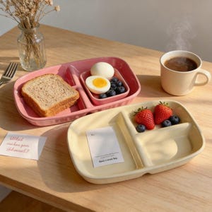 Peut inclure: Deux assiettes en céramique divisées, une rose et une crème, contenant chacune des aliments. L'assiette rose contient du pain, des œufs et des myrtilles. L'assiette crème contient des fraises et des myrtilles. Une tasse de café et une petite note sont également présentes.