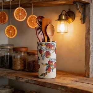 May include: A ceramic utensil holder with a lychee fruit design, holding wooden spoons, a fork, and a spatula. The holder sits on a wooden shelf with glass jars and dried orange slices hanging above. A vintage-style wall sconce provides warm lighting.