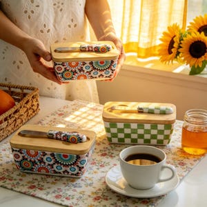 Może przedstawiać: Ceramiczne maselniczki z drewnianymi pokrywkami i pasującymi szpatułkami. Jedna ma kolorowy kwiatowy wzór, druga zielono-biały wzór w kratę. Na zdjęciu znajduje się również filiżanka kawy i słoneczniki.