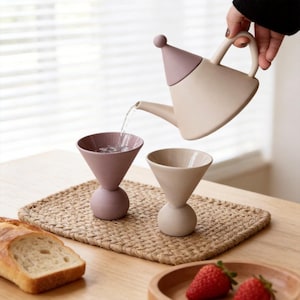 May include: A beige teapot with a pink conical lid pours water into a mauve-colored glass. A second beige glass sits beside the first. The glasses are on a woven mat, with bread and strawberries nearby. The teapot and glasses have a minimalist design.