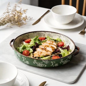 May include: An oval, green ceramic serving dish with a floral pattern, filled with a salad of lettuce, tomatoes, and grilled chicken. The dish has brown handles and a decorative rim. A white bowl and plate are in the background.