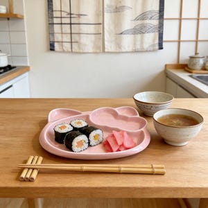 May include: A pink, bear-shaped plate holds sushi rolls with salmon and pickled ginger. Two small ceramic bowls and a pair of bamboo chopsticks rest on a wooden table. The scene suggests a Japanese-themed meal.
