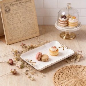 May include: A white rectangular serving tray with gold bow accents holds a small frosted cake and two macarons. A small cake stand with two mini cakes under a glass dome is in the background. An open book and dried flowers are also present.