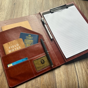 Può includere: Portfolio in pelle marrone aperto con più tasche contenenti un passaporto, un taccuino Field Notes, carte di credito e una carta American Express. Sono visibili anche una penna e un blocco note con una clip.
