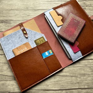 May include: Open brown leather journal with multiple pockets, holding a small red book, cards, and a gray felt insert. The journal is open on a wooden surface, showcasing its interior design and storage options.