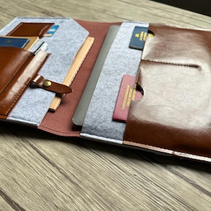 Op de afbeelding: Bruin lederen en grijs vilten reisorganizer met meerdere vakken. Voorzien van een pennenhouder, paspoortvakken en een sectie voor een tablet of notitieboek. Inclusief een drukknoopsluiting.