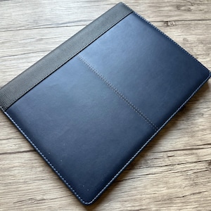 May include: A navy blue leather portfolio with a textured grey spine. The portfolio has stitched detailing and a smooth surface. The item is shown on a wooden surface.