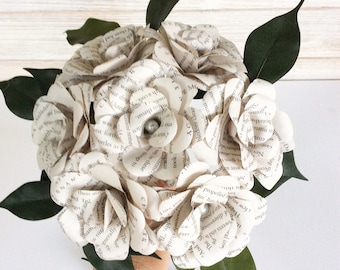 May include: A bouquet of white paper flowers made from book pages. The flowers are arranged in a circular shape and have a pearl in the center. The bouquet is held by a hand.
